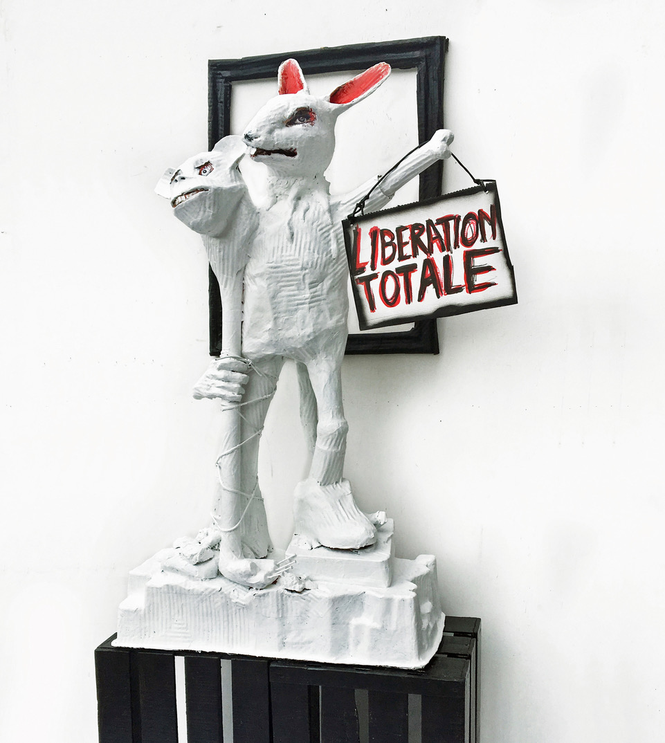 White two-headed animal-like sculpture holding a sign reading “Libération Totale.”
