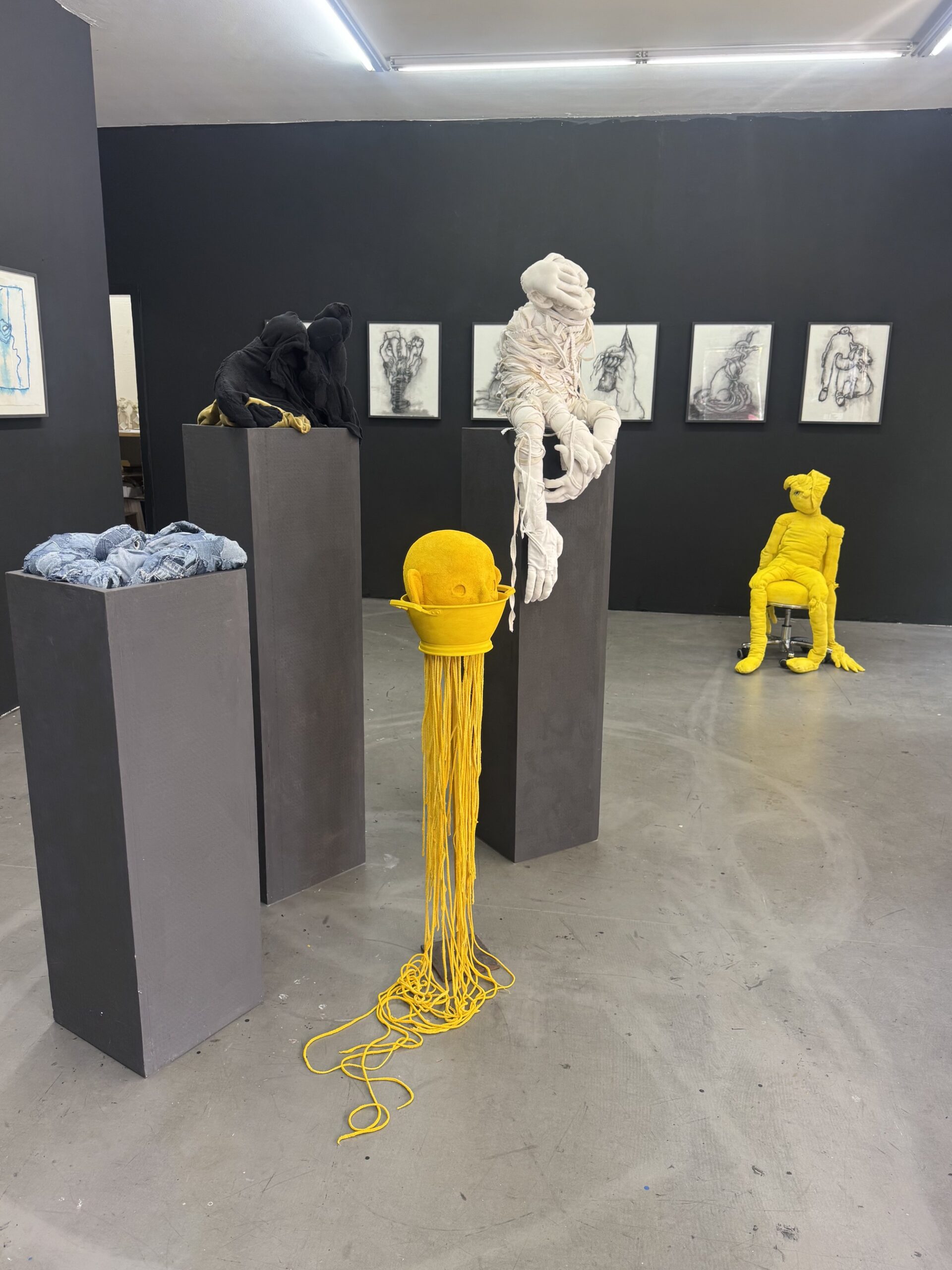 Installation view at Galerie Daeppen, Basel with textile sculptures on dark plinths, including black, white and yellow figures, with framed drawings on the dark gallery walls.