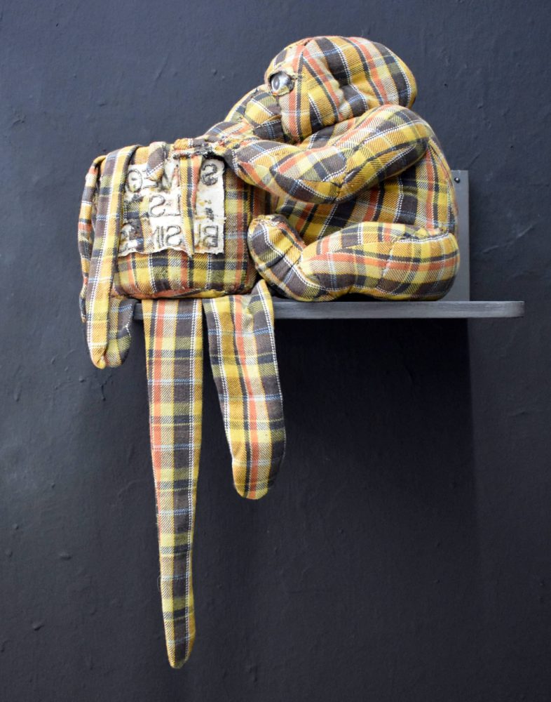 Detail view at Galerie Daeppen, Basel, of an entangled figure with its nose stuck in a pot, made from yellow and brown plaid textile and seated on a black wall-mounted shelf.
