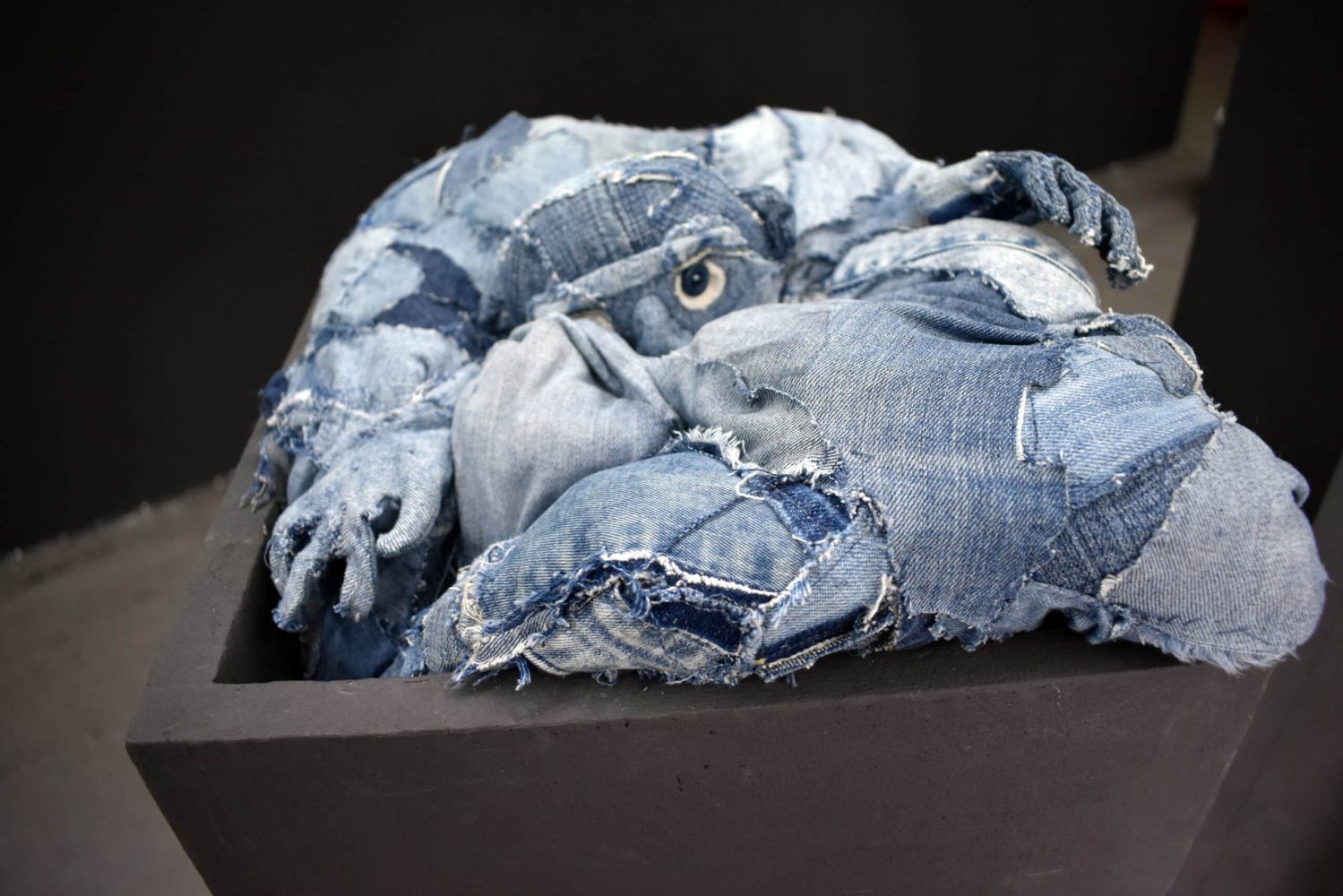Detail view at Galerie Daeppen, Basel, of a denim textile figure sinking into, or trying to climb out of, a dark pedestal.