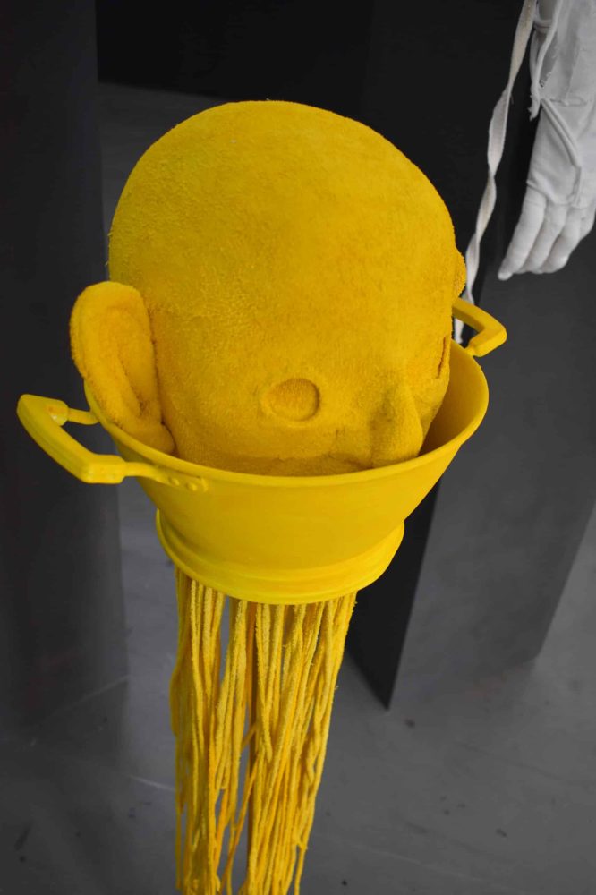 Detail view at Galerie Daeppen, Basel, of a yellow head made of sponge fabric resting in a colander, with long thread spilling onto the floor.