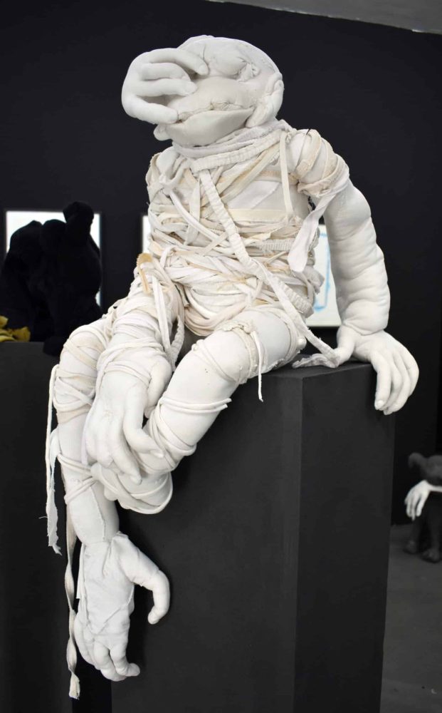 Detail view at Galerie Daeppen, Basel, of a textile figure wrapped and entangled in strips of fabric, seated on a dark plinth with elongated hands hanging down.