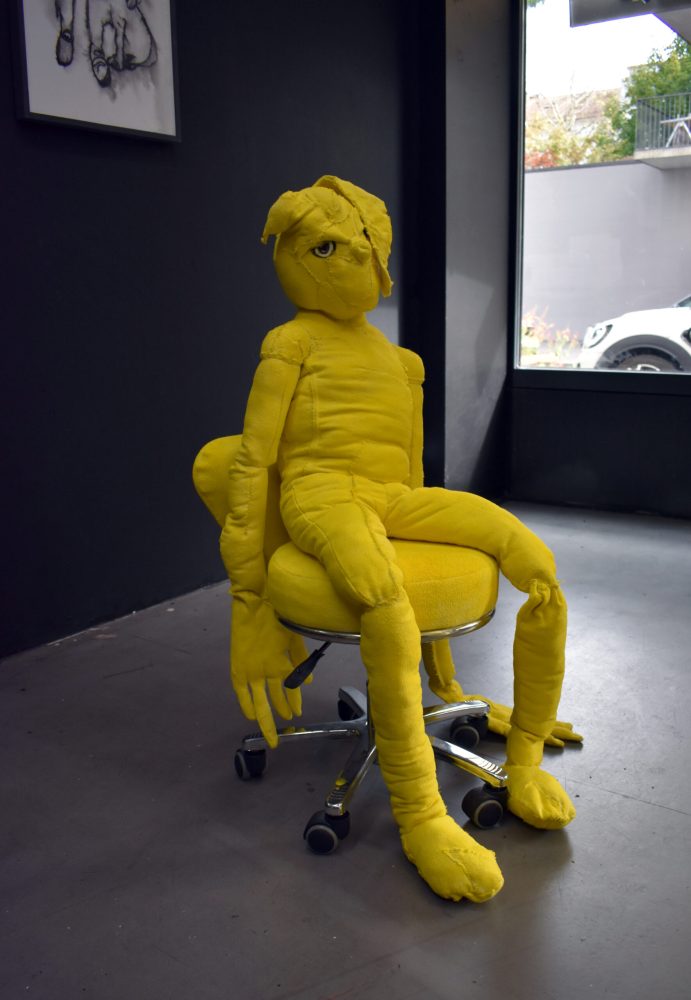 Yellow textile figure seated on a small wheeled stool against a dark gallery wall.