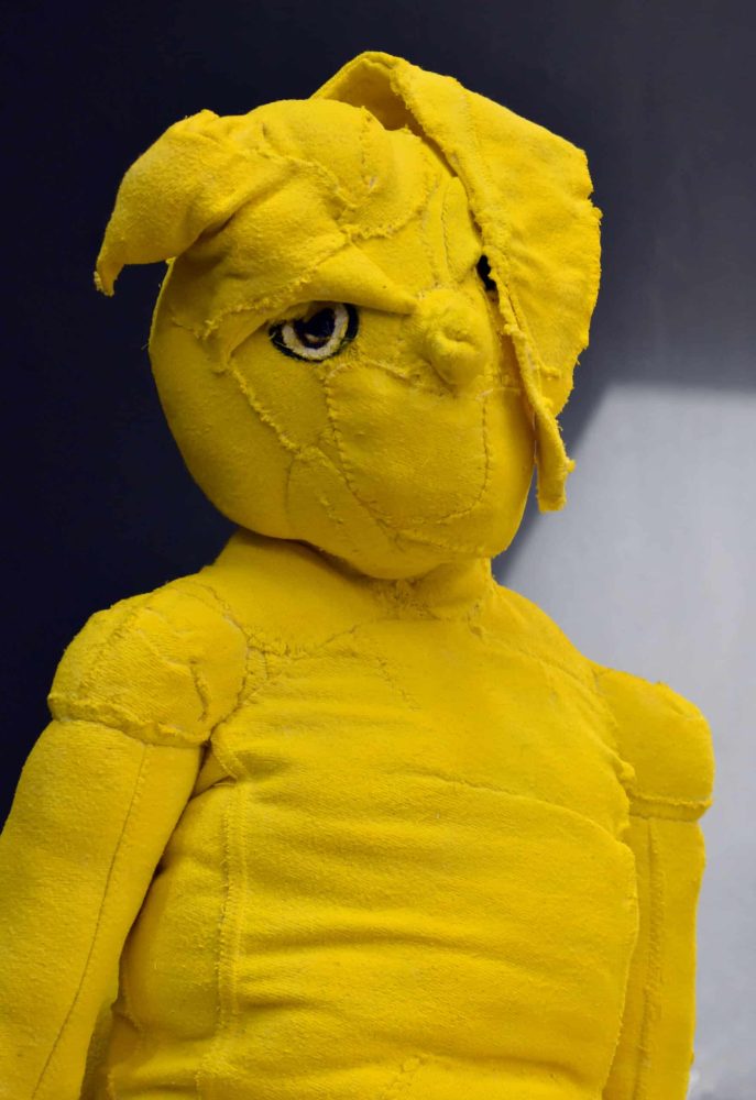 Yellow textile figure seated on a small wheeled stool against a dark gallery wall.