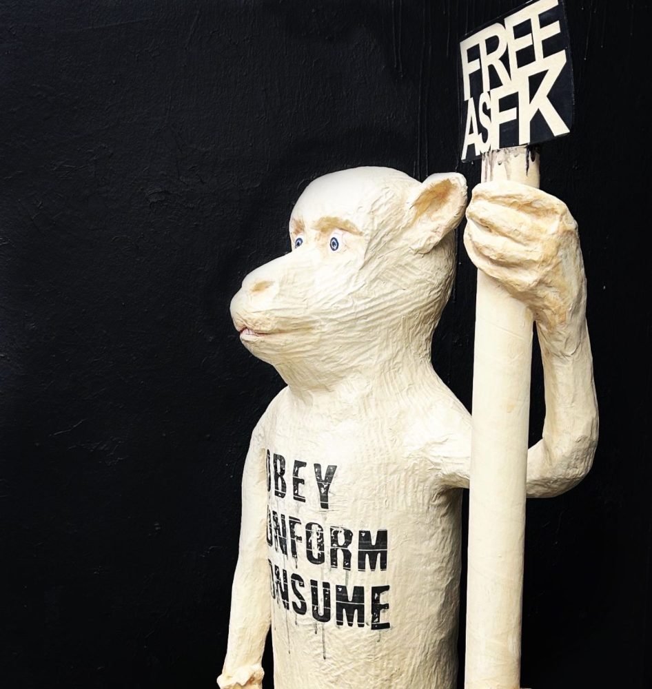 Detail of OBEY CONFORM CONSUME, showing the figure holding a sign reading “FREE AS FCK”.