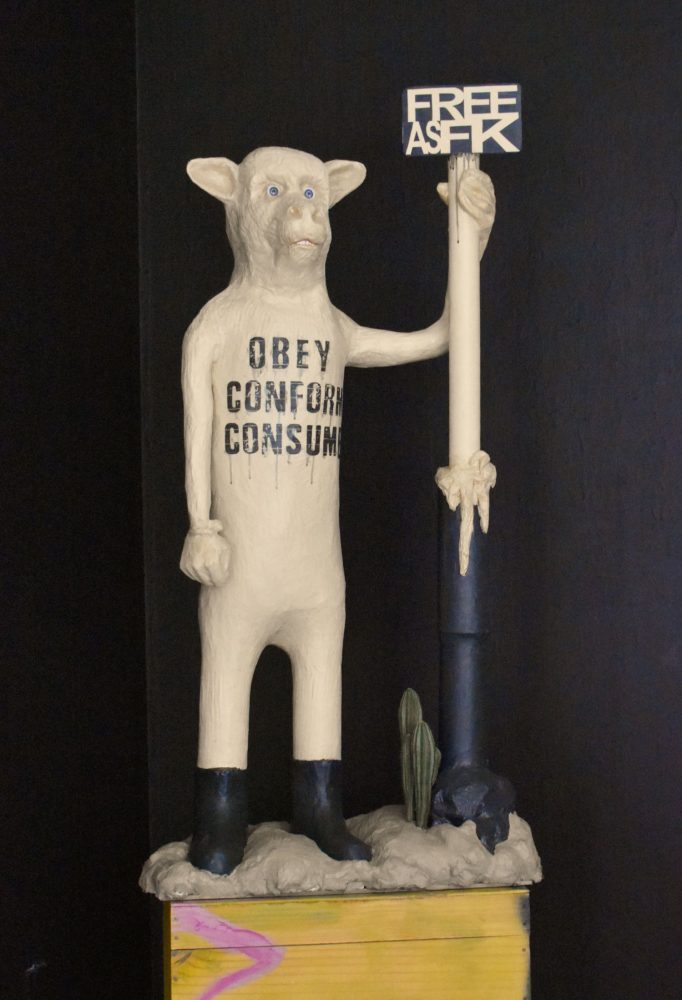 Standing sculptural figure with “OBEY CONFORM CONSUME” on its torso, holding a sign reading “FREE AS FCK”.