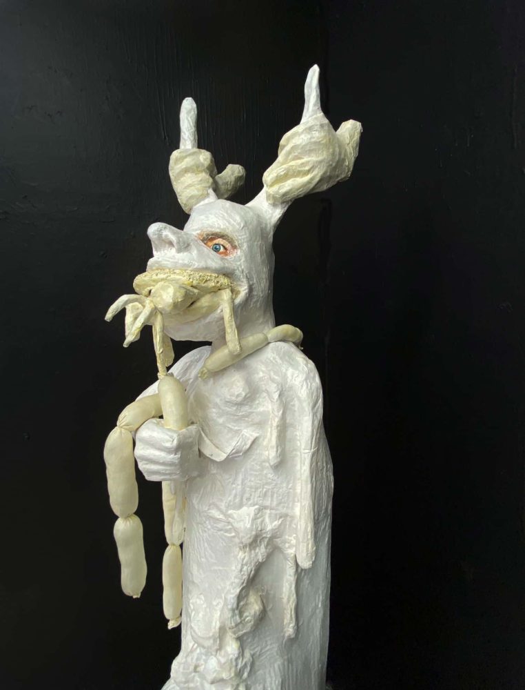 Detail of IST MIR WURST, showing two external hands gripping the horns and a hamburger with animal parts in the figure’s mouth.