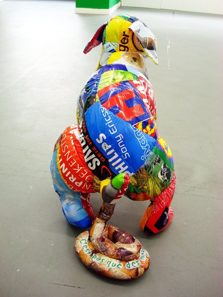 Colourful dog-like sculptural figure defecating, with the words “C’est pas que des mots” written on the excrement.
