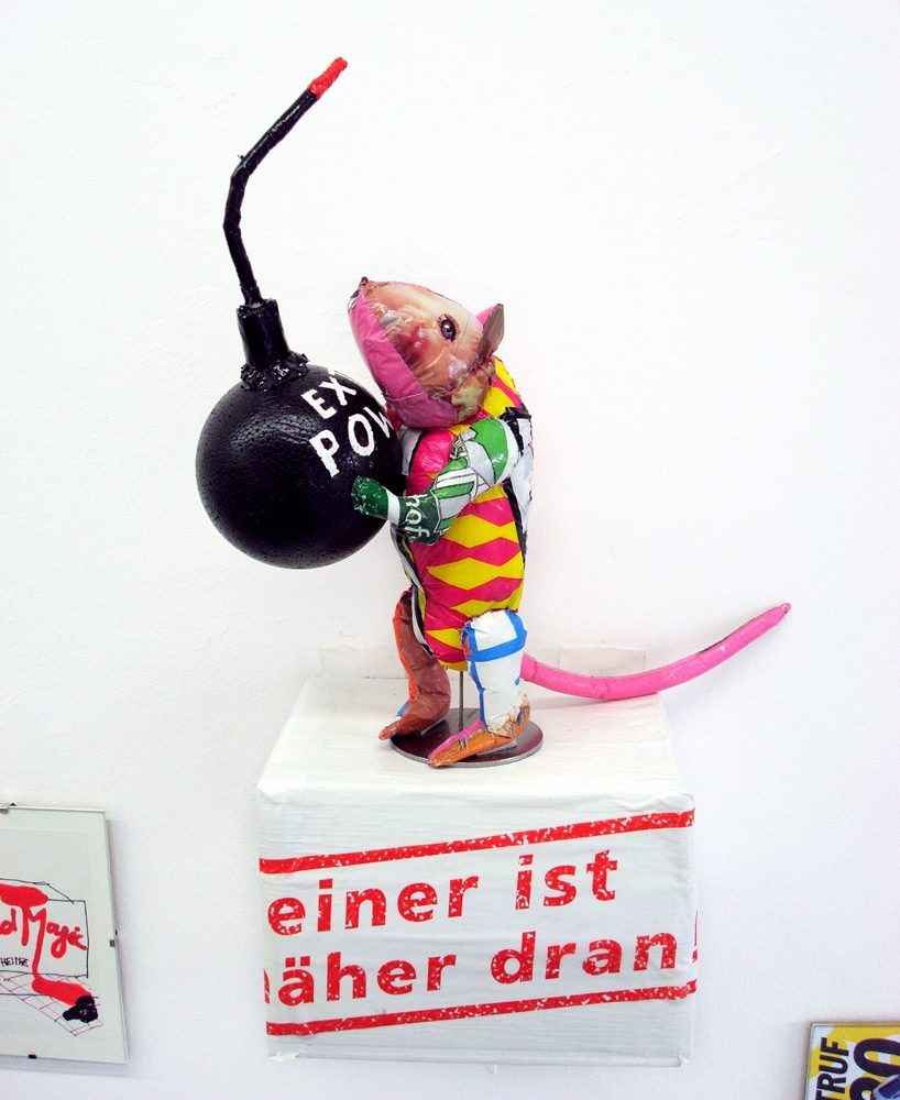 Rat sculpture made from plastic bags holding a bomb, installed on the wall-mounted wooden box Eine ist eher dran.
