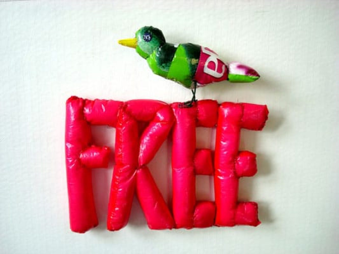Wall-mounted sculptural work made from plastic bags, forming the word “FREE”, with a small green bird-like figure perched on top.