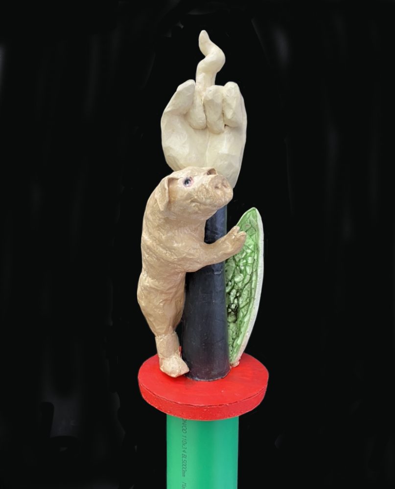 Piglet holding a hand with middle finger in the shape op a pig tail- mounted on a standing green PVC pipe
