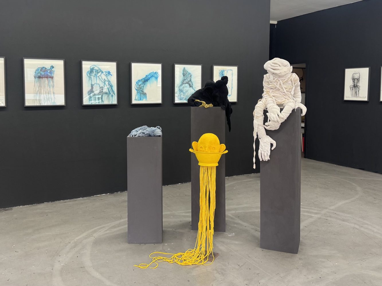 Installation view at Galerie Daeppen, Basel, featuring textile figures in white, black and yellow on dark plinths, with a row of framed blue watercolor drawings.