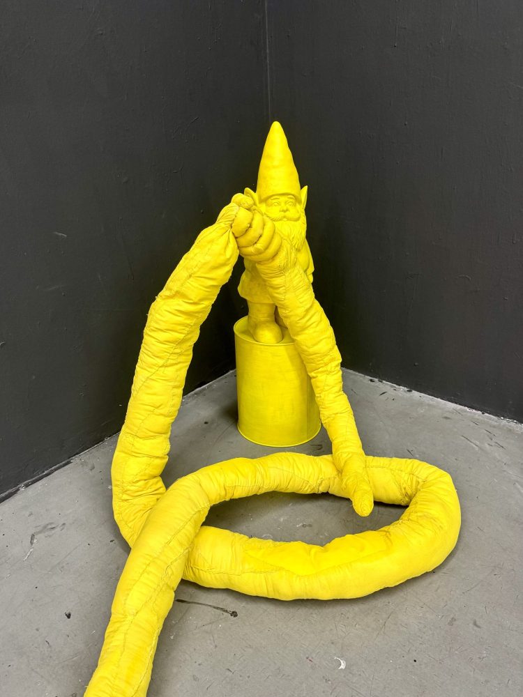 Yellow sculptural figure with elongated arms extending across the floor toward a seated gnome-like figure.