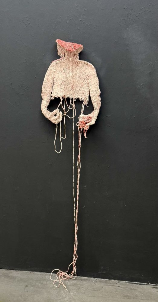 Detail view at Galerie Daeppen, Basel, of a pink knitted, painted and hardened textile figure, with one hand unraveling the torso and the other holding loose threads, while long strands trail to the floor.