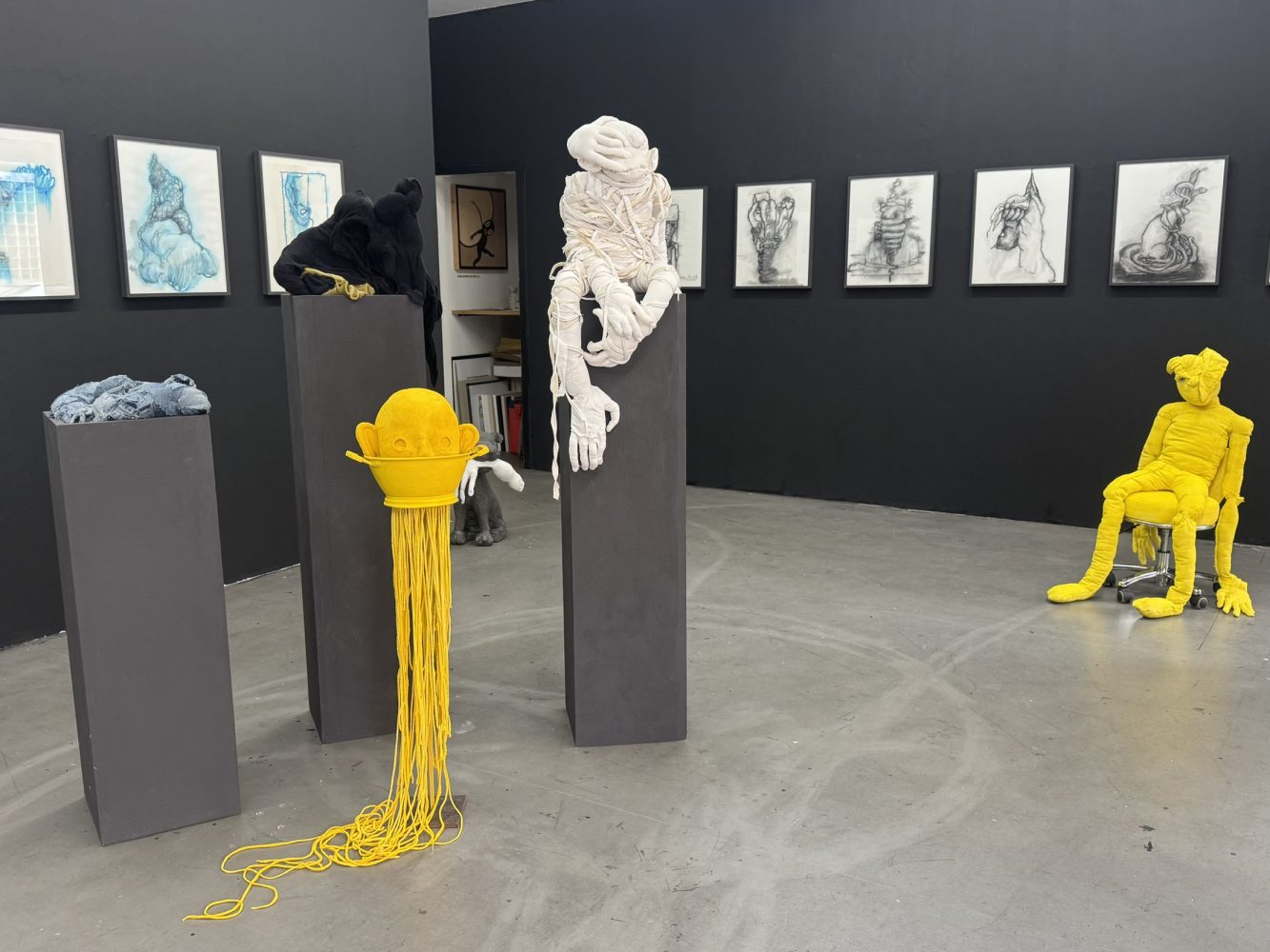 Installation view at Galerie Daeppen, Basel with textile sculptures on dark plinths, including black, white and yellow figures, with framed drawings on the dark gallery walls.