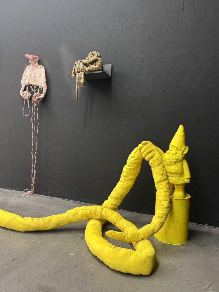 Installation view at Galerie Daeppen, Basel, featuring a yellow garden gnome standing on a yellow metal barrel, entwined with an elongated yellow textile figure, with pink, knitted, hardened work on the dark wall.
