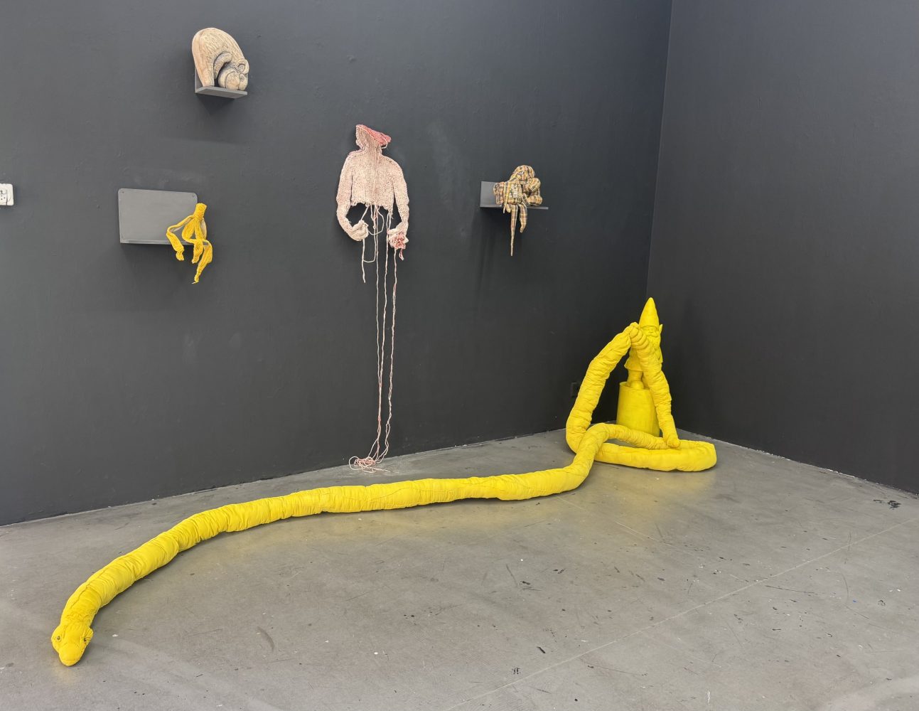 Installation view at Galerie Daeppen, Basel, featuring a long yellow textile figure extending across the floor, with smaller textile works displayed on the dark gallery wall.