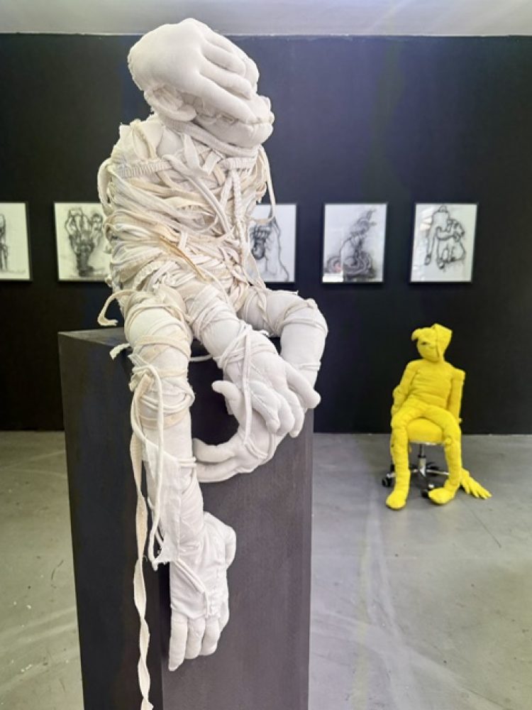 Installation view with a white bound textile figure on a pedestal, a seated yellow figure and framed drawings on a dark wall.
