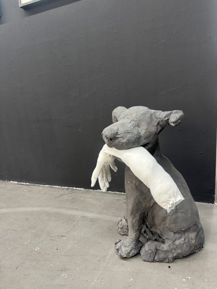 Detail view at Galerie Daeppen, Basel, of a seated dark grey painted papier-mâché figure holding a long white fabric arm and hand in its mouth, sitting on the concrete floor against a dark exhibition wall.