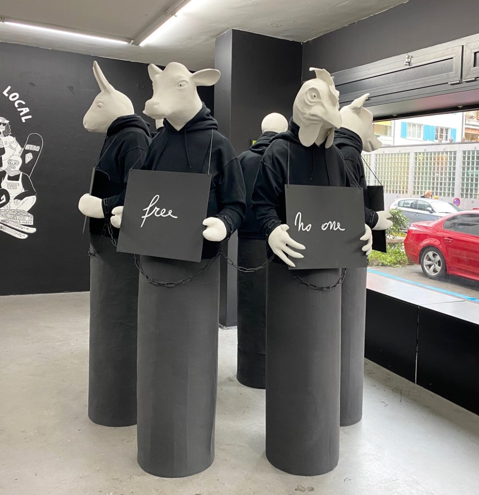 Installation view of five sculptural figures, five with animal heads and one with a human head, holding signs with text fragments, their hands chained together.