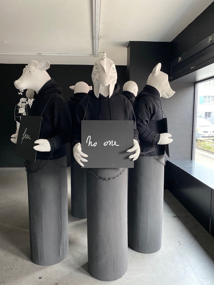 Full view of five sculptural figures on cylindrical plinths, holding signs with text fragments, their hands linked together by chains.
