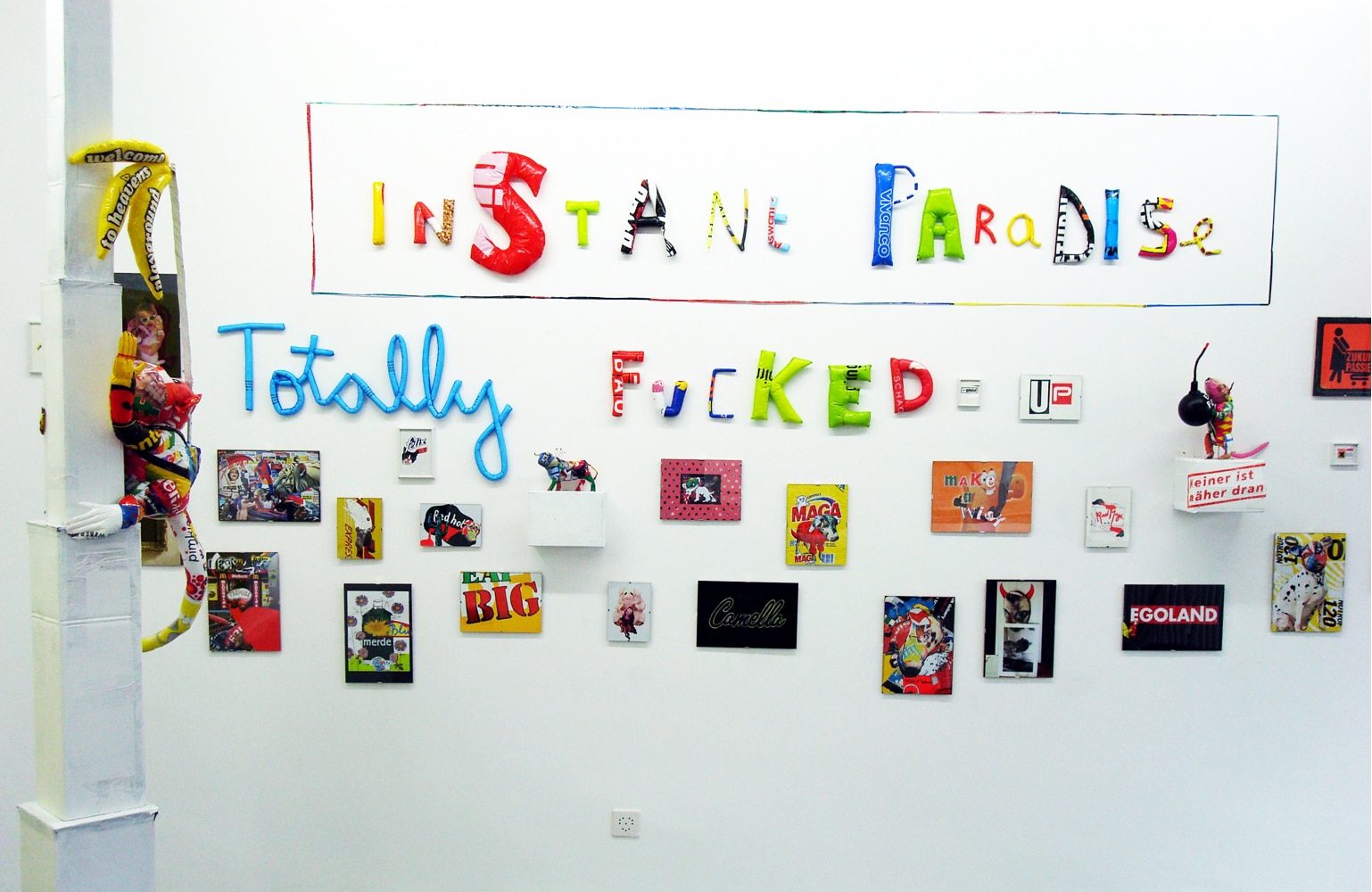Installation wall with the words “INSTANT PARADISE / TOTALLY FUCKED” and a group of small works.