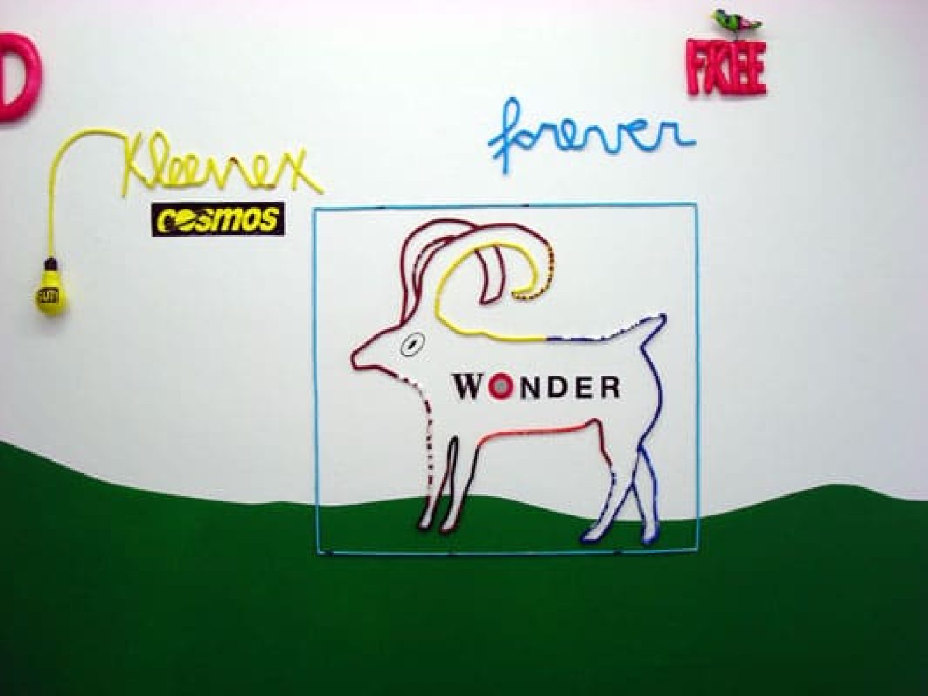 Detail of a wall installation with the words “Kleenex cosmos and forever FREE” and an animal-like figure formed from plastic-bag cord.