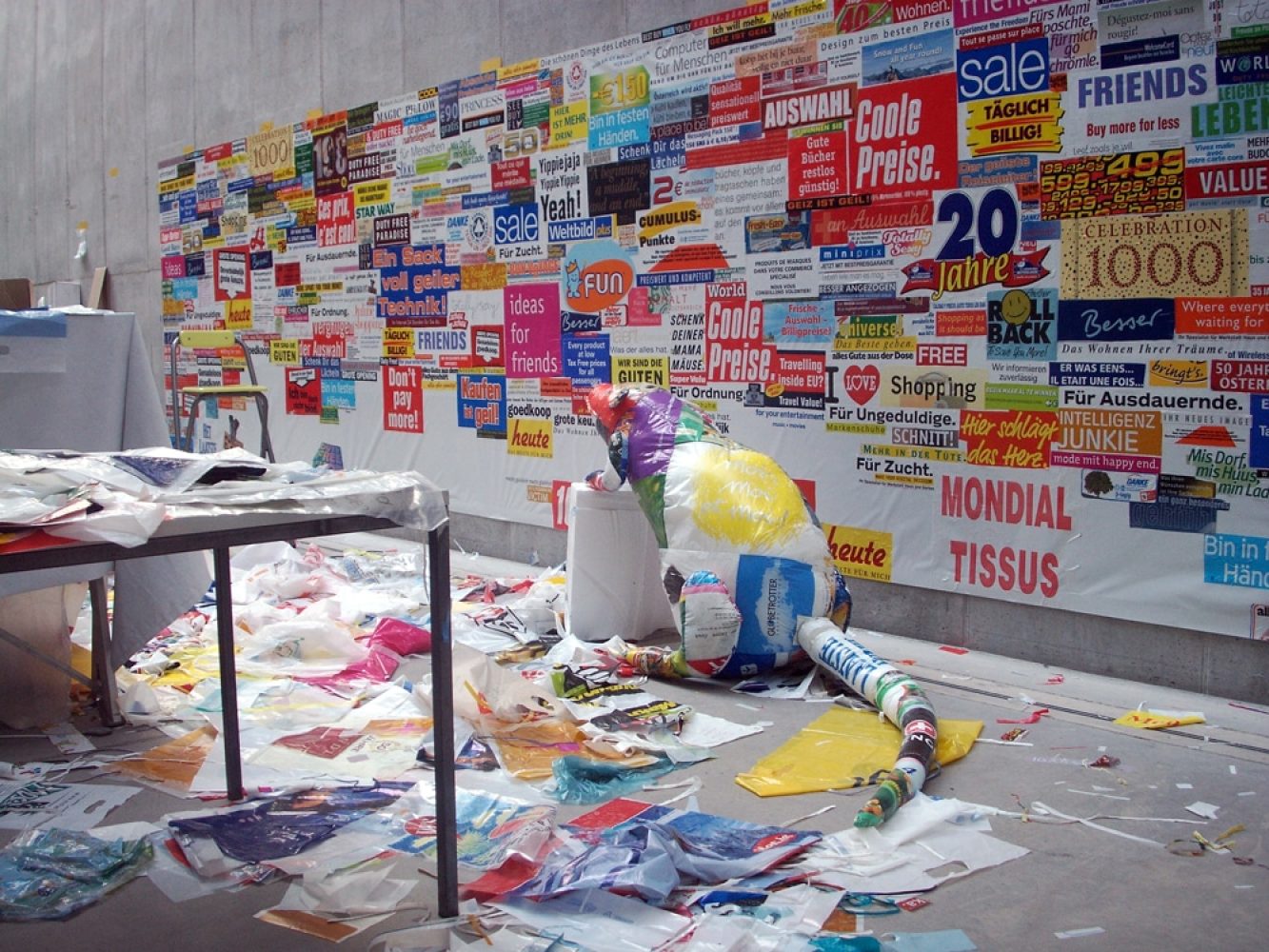Studio installation with a rat-like sculptural figure, advertising material and a large accumulation of discarded plastic bags.
