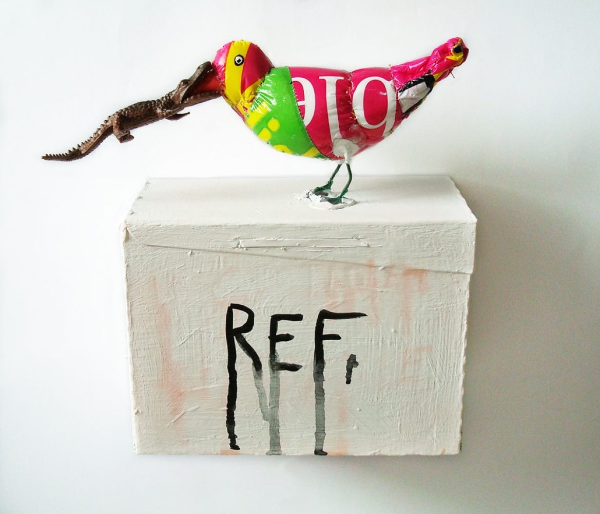 Bird made from a plastic bag holding a toy crocodile in its beak, perched on a white wooden box marked “REF”.