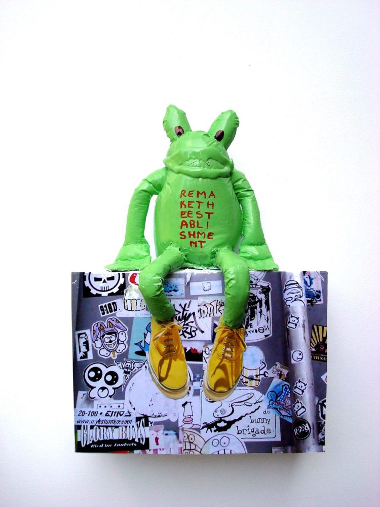 Green frog-like figure made from painted plastic, seated on a collage-covered wooden base.