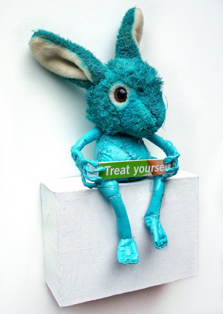 Blue rabbit-like figure made of painted plastic and plush, seated on a white wooden box with a small text plaque.