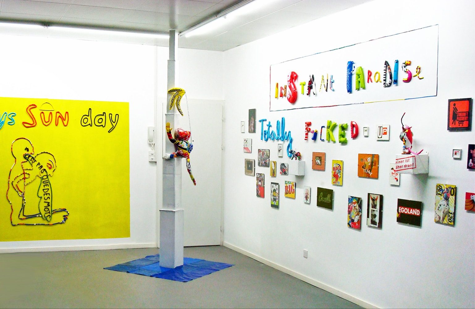 Installation view with the words “INSTANT PARADISE / TOTALLY FUCKED” on the wall and a sculptural monkey climbing toward an unreachable bunch of bananas.