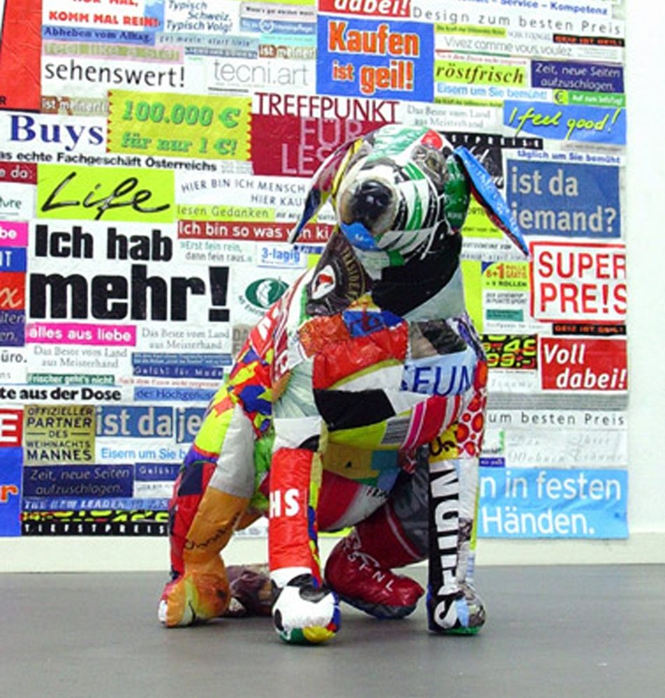 Alt Text Installation view of a defecating dog sculpture made from printed plastic bags in front of a wall collage of advertising messages cut from plastic bags.