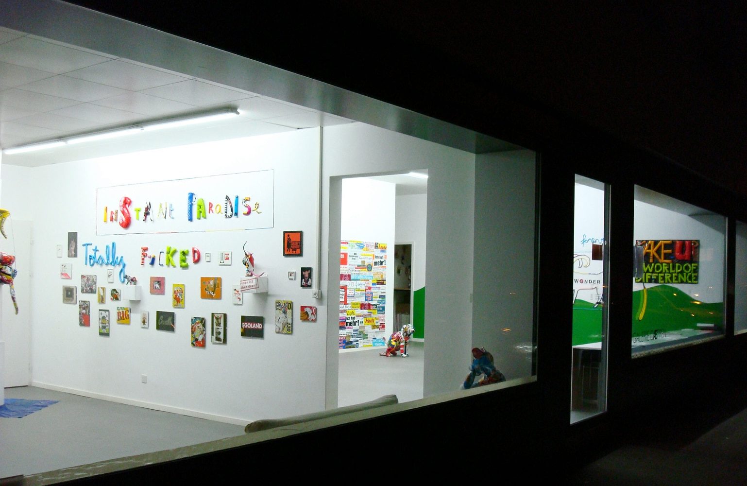 Exterior view through the gallery window showing the installation with wall works and sculptural figures inside.