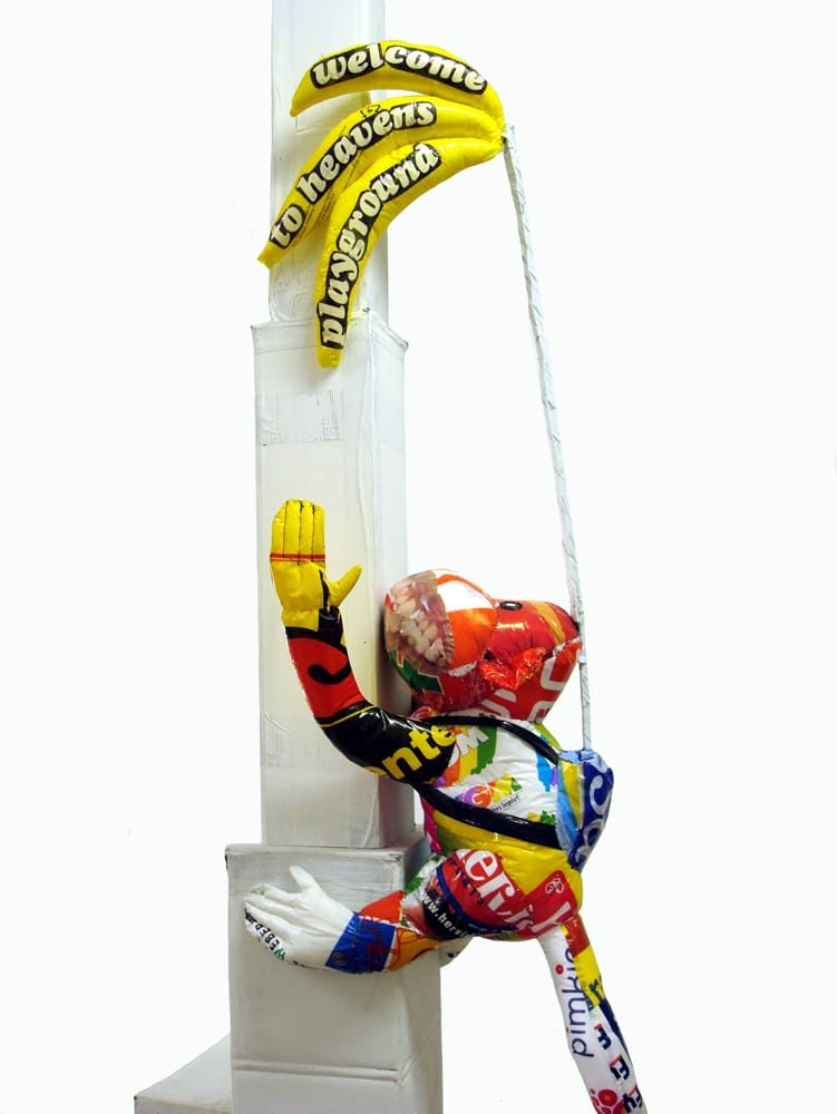 Sculptural monkey made from plastic bags, climbing upward with a stick attached to its back holding a bunch of bananas permanently out of reach.