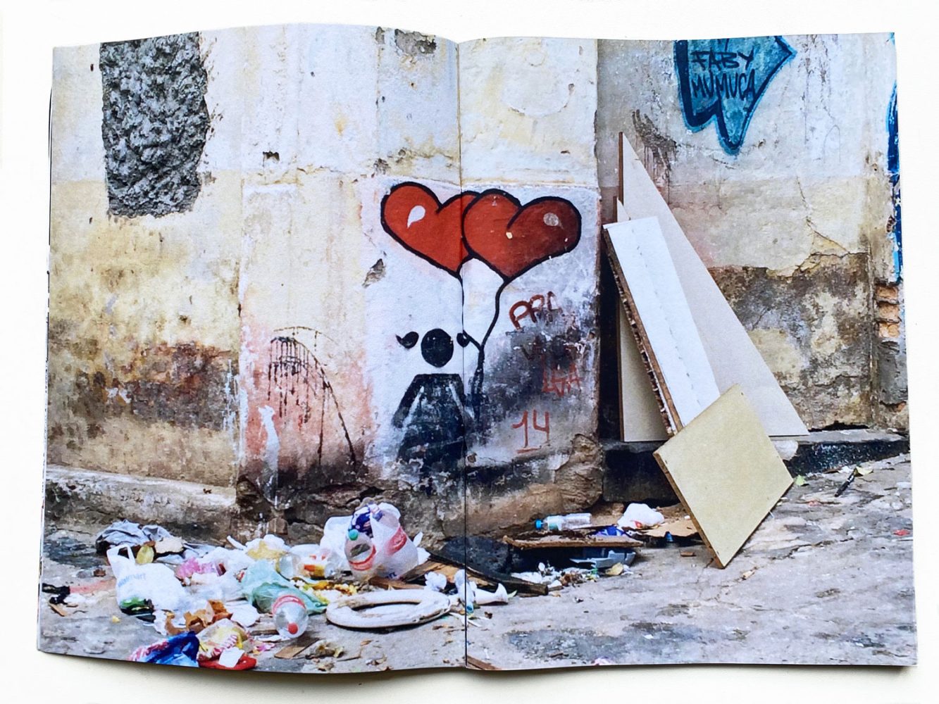 Discarded objects and debris beside a weathered wall with a girl with red heart balloons graffiti in São Paulo.