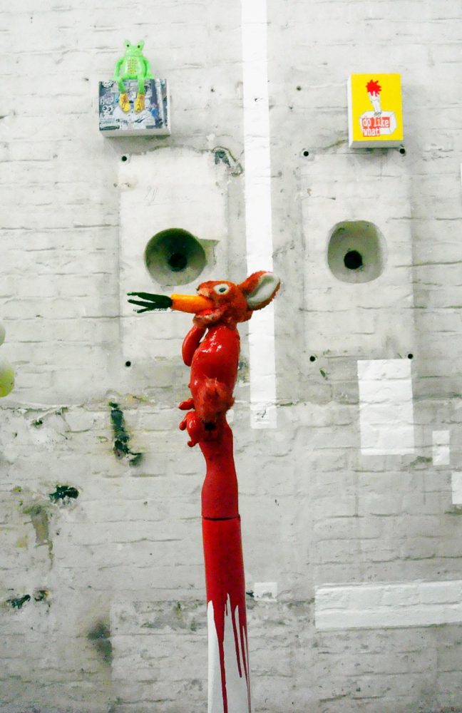 Installation view with a red elongated figure and smaller sculptural works displayed on a pale wall.