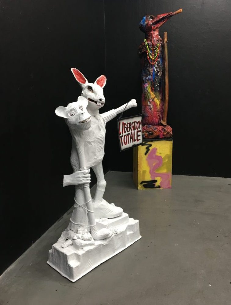 Installation view of Libération Totale, a white two-headed animal-like sculpture holding a sign, at Sweet Bunny Cute Bunny, Basel.