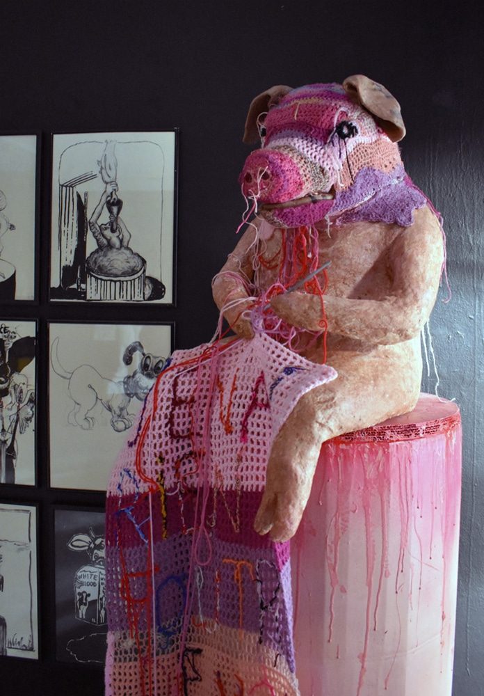 Detail of Rien à foutre, showing a papier-mâché pig crocheting a textile that reads “J’en ai rien à foutre de vos traditions.”