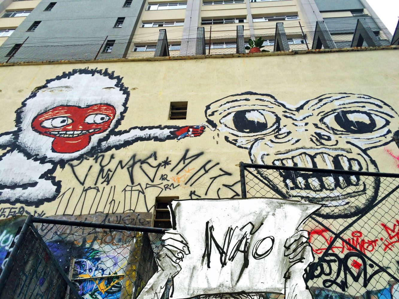 Photomontage of a graffiti-covered wall in São Paulo with two hands holding a sheet reading “NÃO”.