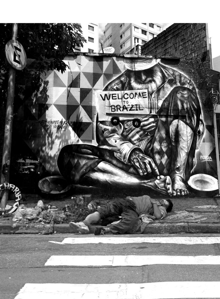 Black-and-white photograph of a person lying on a São Paulo sidewalk in front of a mural with the words “Welcome to Brazil”.